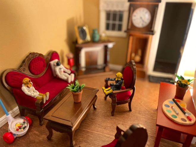 Miniature astronaut and scuba divers sitting on miniature furniture talking with a grandfather clock and fireplace behind them, a paint palette, and toy