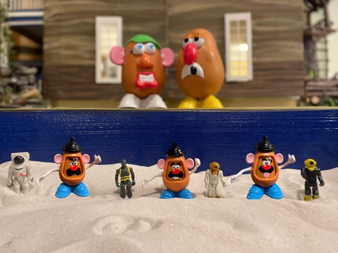 Same toys from last pictures together with 5 Potato Head toys with different facial expressions standing in a sand tray in front of a dollhouse