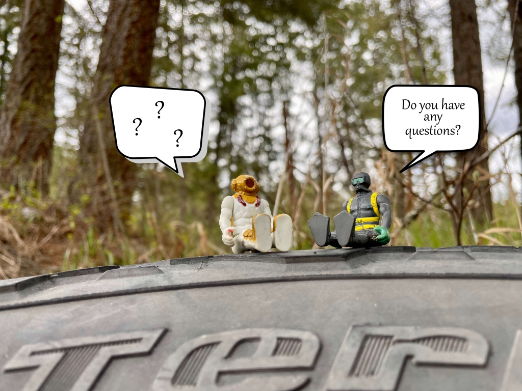 Toys sitting on a tire out in nature. One toy is asking the other if they have any questions and the other has a talking bubble filled with questions marks