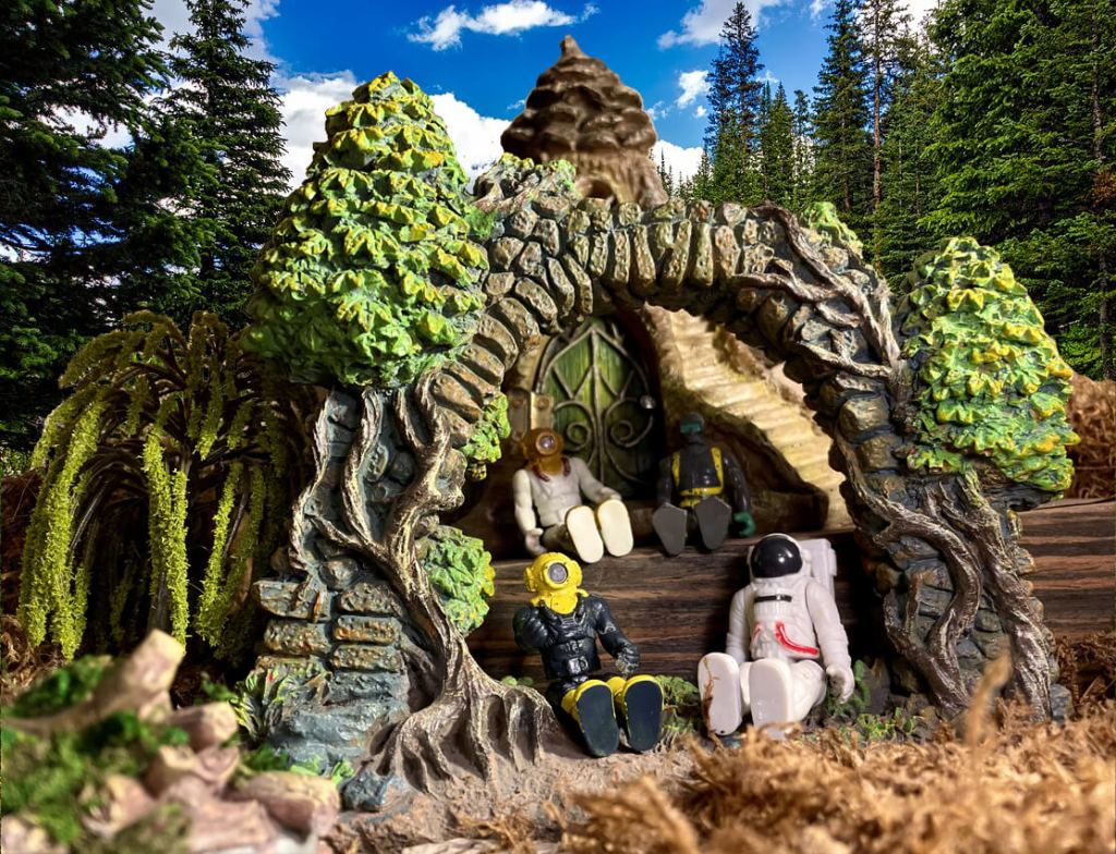 4 toys sitting under an arch made of trees and bricks in front of a cabin with a green door and pinecone roof in a forest